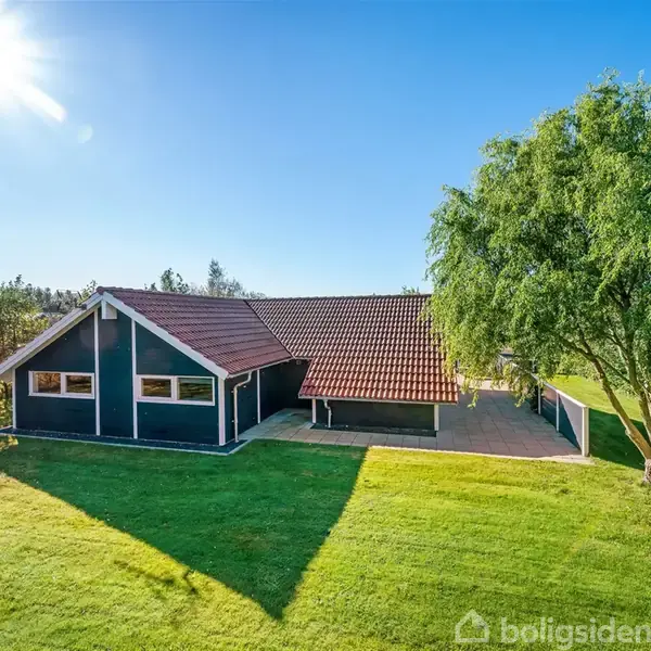 Et sort hus med rødt tag står på en grøn græsplæne. Solen skinner fra en klar blå himmel, omgivet af træer og buske.