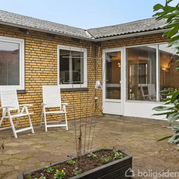 To hvide stole står på en flisebelagt terrasse foran en murstensvæg med vinduer. Omkring er der grønne planter og en åben dør til et indendørs rum.