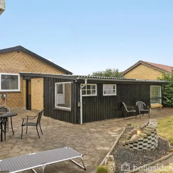 En møbleret terrasse med bord og stole står foran et gult murstenshus og en sort træbygning. En bålplads er omgivet af græs og buske i baggrunden.