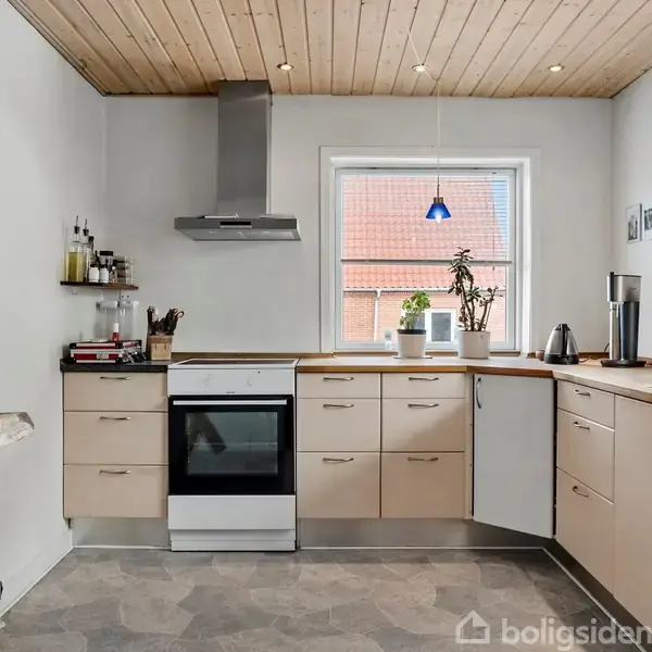 Køkken med hvide skabe og stålgreb står stille. En ovn og kogeplade er centreret, mens et vindue med udsigt til et tag bagved giver lys.