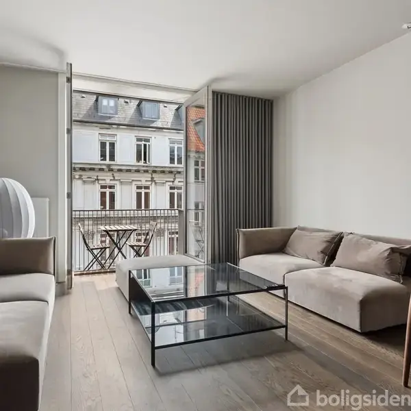 To sofaer står stille i en stue med trælaminatgulv, store vinduer og en balkon med udsigt over en bygning. Et glas-sofabord og rund lampe kompletterer indretningen.