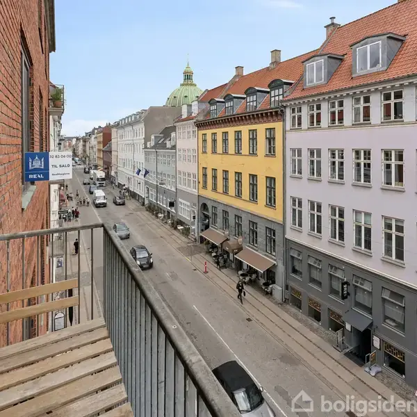 En smal balkon med træstole hænger over en gade med biler og farverige bygninger. I baggrunden ses en kuppel. Et blåt skilt på muren siger “Velkommen” og “PNB A/S”.