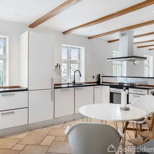 Et moderne køkken med hvide skabe i en åben planløsning. Rundt bord med stole står midt i rummet. Store vinduer giver naturligt lys, træbjælker ses i loftet.