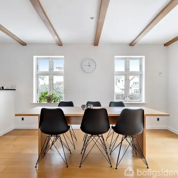 Et spisebord med seks sorte stole står i et lyst rum med trægulv og synlige loftsbjælker. To vinduer og en vægur pryder væggen bagved.