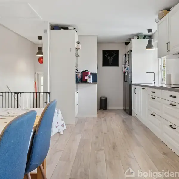 Et køkken med hvide skabe og træbordplader. Et spisebord med blå stole står i forgrunden. Rummet har lyse trægulve og vægophængte lamper i en moderne stil.