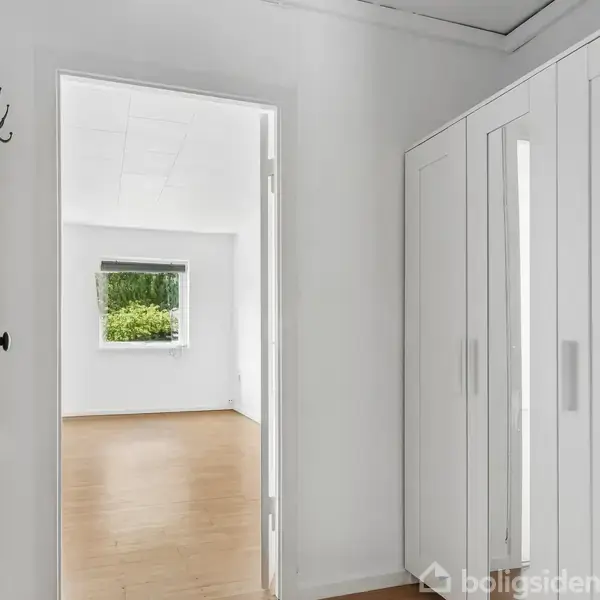 En hvid garderobe står i en lys gang med trægulv. En dør åbner ud til et værelse med vindue, der viser grønne træer udenfor.