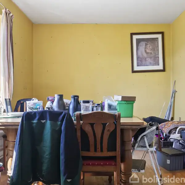 Et bord er dækket med diverse genstande som flasker og kasser. Rummet har gule vægge, et stort vindue med gardiner, og en stol ved bordet. En indrammet billed hænger på væggen.