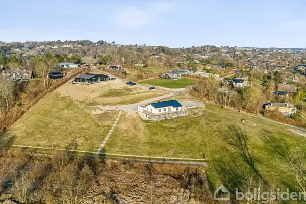 En moderne, lav bygning ligger på en stor, græsbevokset grund på en bakke. Omkring bygningen ses en landlig bebyggelse med flere huse og træer i baggrunden under en klar himmel.
