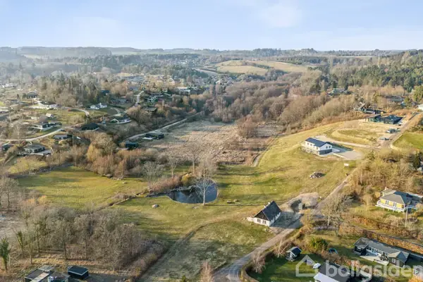 Landområde med spredte huse, omgivet af bakker og skov. En vej snor sig gennem landskabet, og vegetation er dominerende. Udsigten strækker sig over et bakket, kuperet terræn i det fjerne.