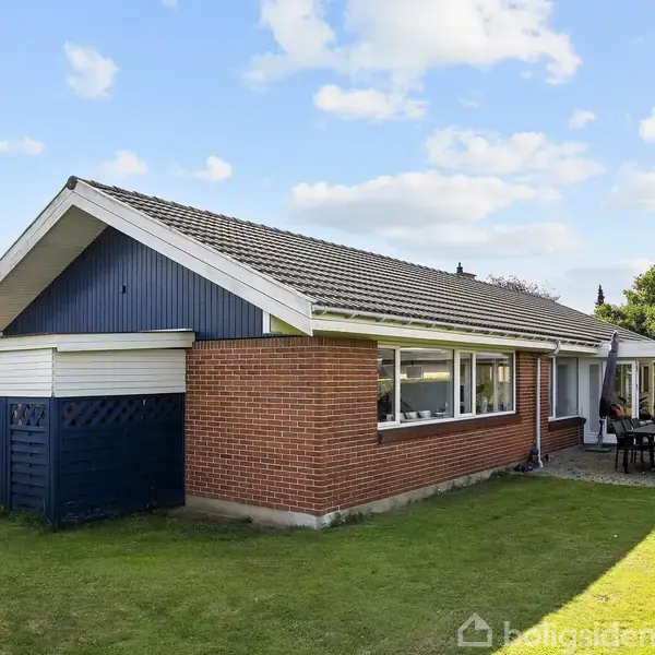 En murstenshus står i en velplejet have. Ved siden af huset er en terrasse med bord og stole, omgivet af grønne træer og en klar blå himmel.