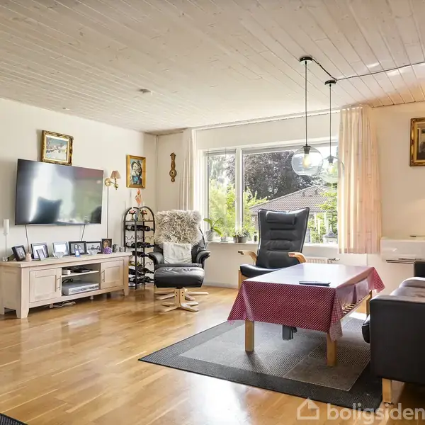 Stue med sofa, lænestol, og sofabord med dug. TV hænger på væggen over en skænk med billeder. Vindue med udsigt til have. Malerier og lamper dekorerer rummet.