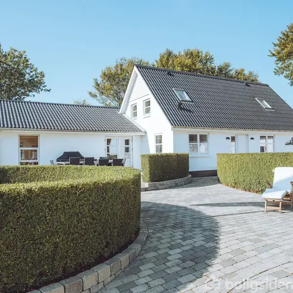 Et hvidt hus med sort tag står i en anlagt have med brosten og klippede buske. En solstol er placeret på terrassen. Træer omgiver området under en klar himmel.