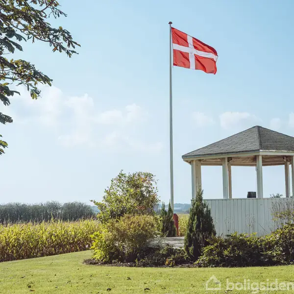 Pavillon står frit på en græsplæne omgivet af buske, med et flagstang ved siden af, hvor Dannebrog vajer. I baggrunden ses en mark og træer.