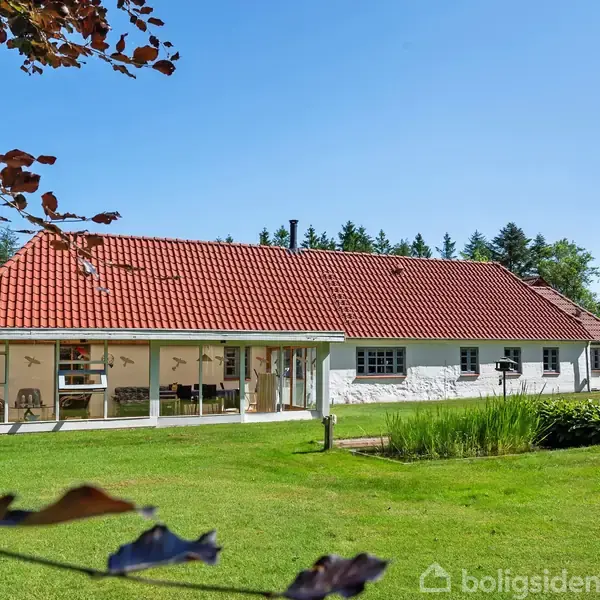 Hus med rødt tegltag står stille i en grøn have. Lyst overdækket område ved huset, omgivet af træer og med en grøn græsplæne. Ingen tekst.