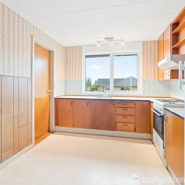 Køkken med træskabe og -skuffer, står i et lyst rum med vindue og dør. Tapet og træpaneler pryder væggene, gulvet er lyst. Fjern miljø med boligblokke udenfor.