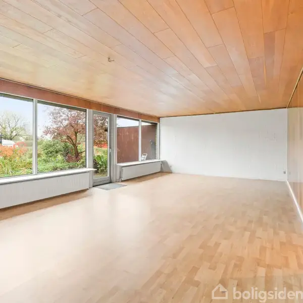 Stue med trægulv og træloft, store vinduer med udsigt til en have og en åben dør, der fører ind i rummet.