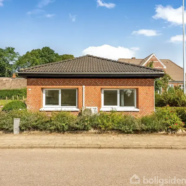 Et lavt rødt murstenshus med to store vinduer mod en vej. Huset er omgivet af en lille have, og i baggrunden ses flere træer og huse under en blå himmel.
