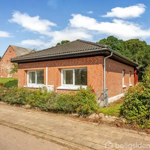 Rødt murstenshus med sort tag står stille på villavej, omgivet af grønne buske og træer. Flere huse er synlige i baggrunden under en blå himmel med få skyer.