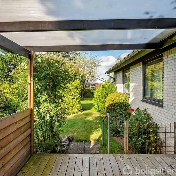 En terrasse med trægulv er dækket af et halvtag. Udgangen fører til en have med grøn, velplejet græsplæne og buske, omgivet af en hvid murstensvæg.