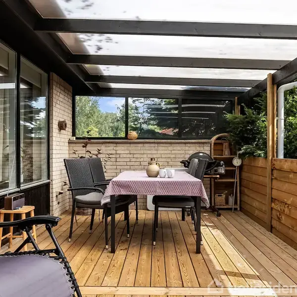 En overdækket terrasse med et bord dækket med dug og en vase. To stole står ved bordet. Der er en liggestol til venstre. Baggrunden omfatter en murstensvæg og grønne planter.