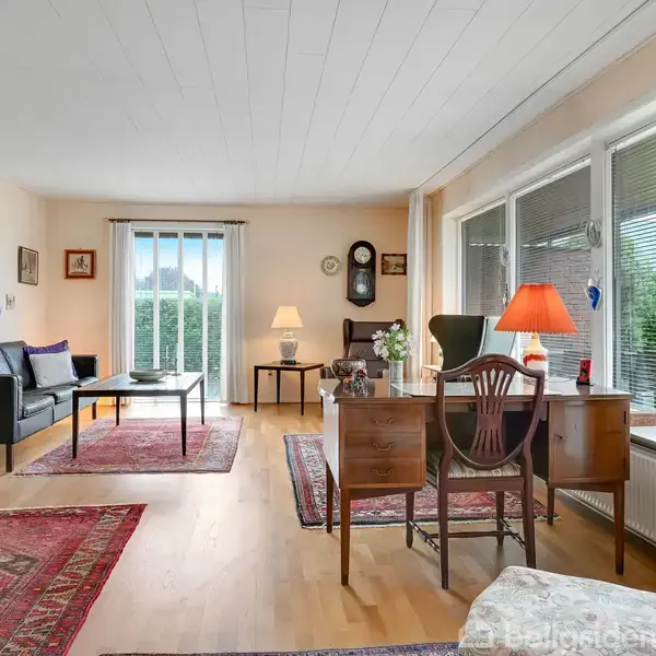 Stue med sofa modsat skrivebord ved vinduet. Rød lampe, tæpper på gulvet og kunst på væggene. Lys strømmer ind fra store vinduer.