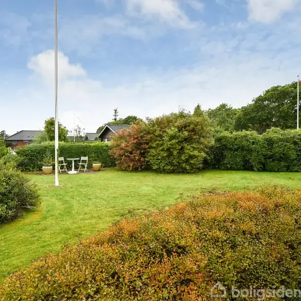 En flagstang står i en grøn have med en tæt omkransning af buske. Et sæt havemøbler er placeret nær flagstangen under en blå himmel.