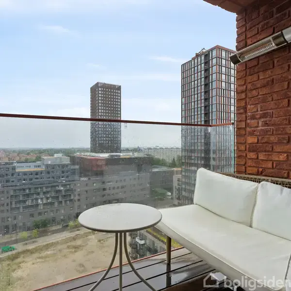 En balkon har en hvid sofa og et rundt bord foran en glasvæg. Baggrunden viser flere høje bygninger i en by med klar himmel.