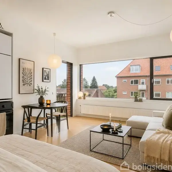 En murstensvæg står stabilt udendørs ved en solrig balkon med sort gelænder. I baggrunden ses tage og træer under en delvist overskyet himmel.