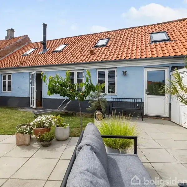 Hus med blå facade og rødt tegltag. En terrasse med grå fliser indeholder en sofa og blomsterkrukker. Græsplæne og hvid dør i baggrunden. Skyfri himmel.