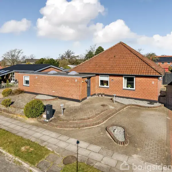 Rødt murstenshus med rødt tag står stille i et parcelhuskvarter. Foran huset er en stenbelagt indkørsel og en lille, enkel have med buske og bede.