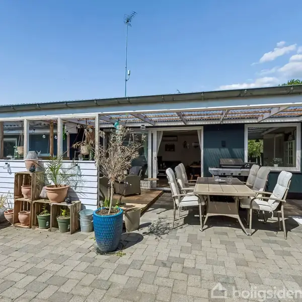 Terrassebord med stole står under overdækket terrasse. Omringet af flere blomsterkrukker og planter i forskellige størrelser. Husets facade er blå med store vinduer, klar himmel i baggrunden.