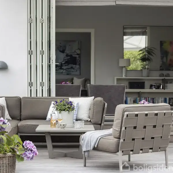 Grå loungesofa med pude og blomster på bordet står udendørs på terrasse, omgivet af kurv med lilla blomster, og i baggrunden ses en indretning med reol og maleri.