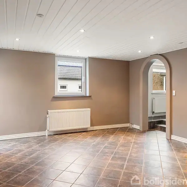 Et tomt rum med brune klinkegulve og hvide trælofter. Dør til venstre fører ud, mens en buegang til højre åbner til et andet rum. En radiator er under vinduet.