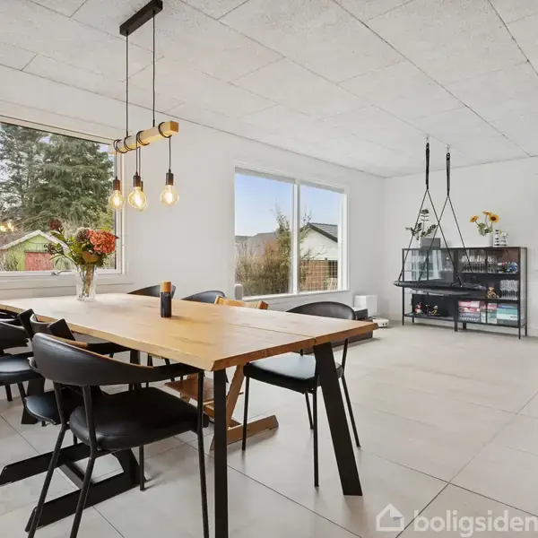 Et elegant spisebord med sorte stole står i et lyst rum. Over bordet hænger moderne pendellamper. Vinduerne giver udsigt til en have. En reol med planter står i hjørnet.