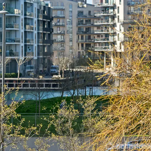 Moderne lejlighedsbygninger står stille ved en kanal, omkranset af træer med spirende blade. Sollys belyser scenen, og en grøn park ligger på den anden side af vandet.
