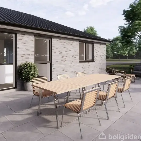 En langbord med stole står på en flisebelagt terrasse. Bagved ses et hus med store vinduer og en åben dør. Træer og en sofagruppe er i baggrunden.