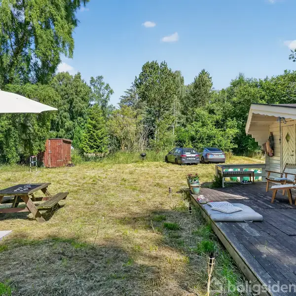 En træterrasse med stole og bord står ved siden af et græsareal med en parasol og picnicbord. To biler er parkeret i baggrunden, omgivet af grønne træer.