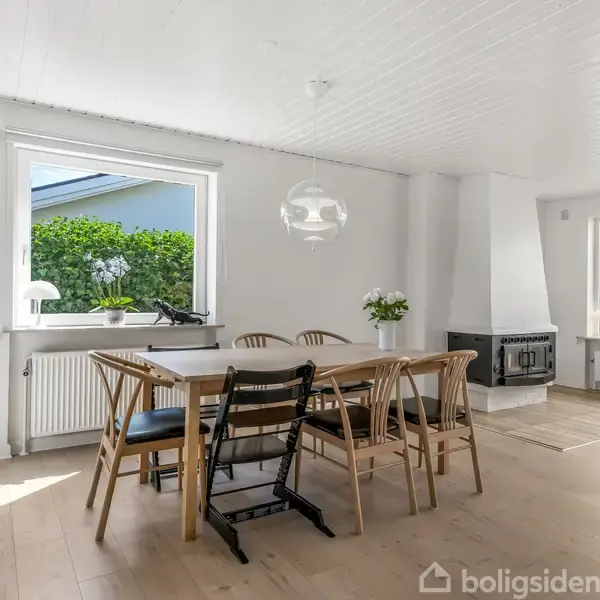En spisestue med træbord og seks stole, hvoraf én er en højstol. Rummet er lyst, med parketgulv, en stor vinduesåbning og en brændeovn i hjørnet.