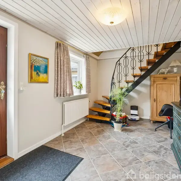 Døråbningsgang med trapper. Trætrappe fører opad, omkranset af planter og møbler. Rummets vægge har kunstmaleri og vindue med gardin, og gulvet er flisebelagt.