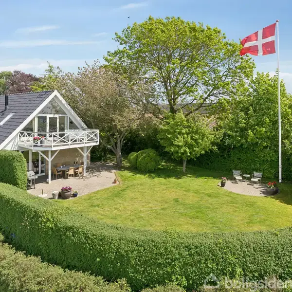 Et gult hus med sort tag har en balkon og terrasse. Det ligger i en velplejet have med en stor flagstang, der bærer et dansk flag.