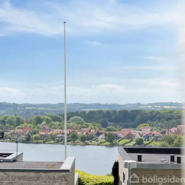 Flagstang står stille med udsigt over en rolig flod, omgivet af moderne bygninger i forgrunden og grønne områder samt huse på den anden bred i baggrunden.