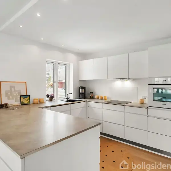 Et moderne køkken med hvide skabe og indbygget ovn. På bordpladen står køkkenredskaber og små dekorationer. Rummet er lyst med vindue, hvidt loft og indbygget belysning.