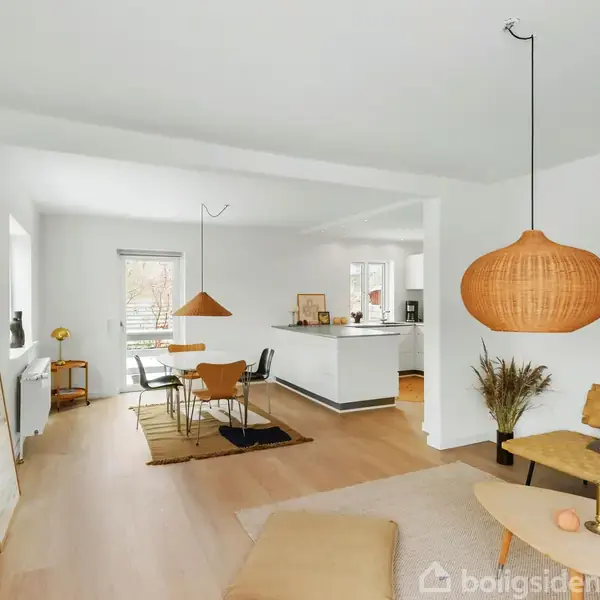 Stue med sofa og sofabord, hængende lamper i træ-look. I baggrunden er der et spisebord med stole. Rummet har store vinduer, der giver naturligt lys, og lyse trægulve.