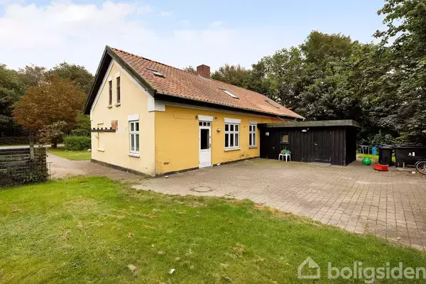 Et gult hus med rødt tegltag står på en brolagt terrasse. Træer omkranser haven i baggrunden, og et sort udhus er ved siden af huset.