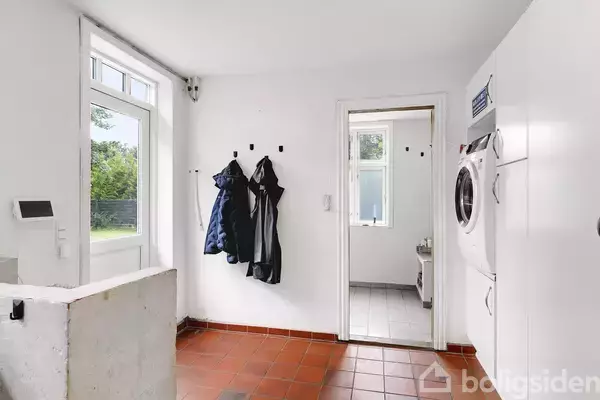 Vaskemaskine hænger på væg; ved siden af dør med jakker hængende under. Rummets flisegulv er rødt, med udsigt til en grøn have.