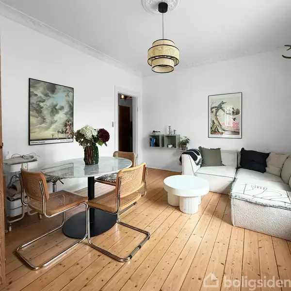 Stuen har et rundt marmorbord med fire stole, buket blomster, og en lys sofa hjørne. Væggene er udsmykket med kunst, og der hænger et gevir. Gulvet er af lyst træ.