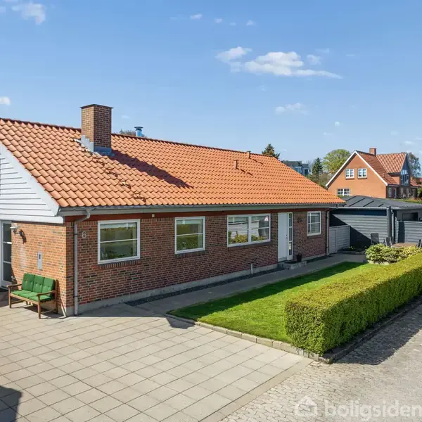 En murstensvilla med rødt tegltag står ved vejen. Den har en lang hæk, flisebelagt indkørsel, og en carport i roligt villakvarter. To grønne bænke står ved husets facade.