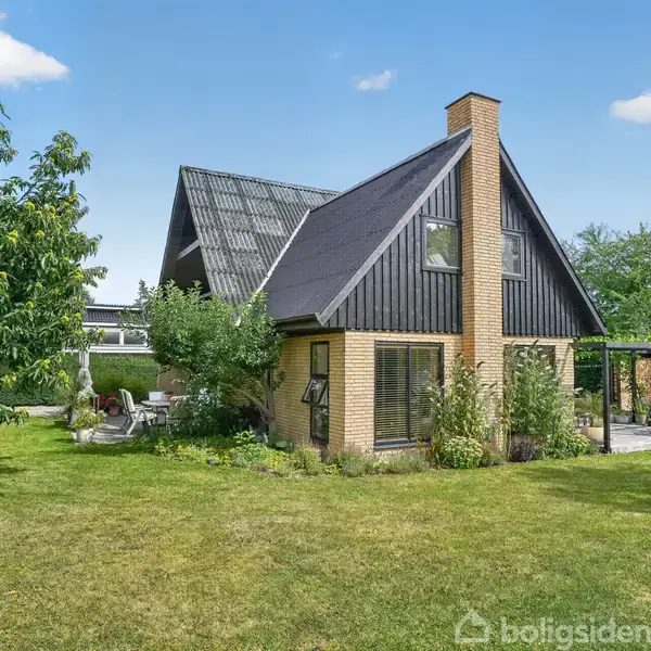 Et hus med skråtag og murstensfacade står i en grøn have med træer og buske. Et terrasselignende område er synligt til venstre, omgivet af græsplæne.