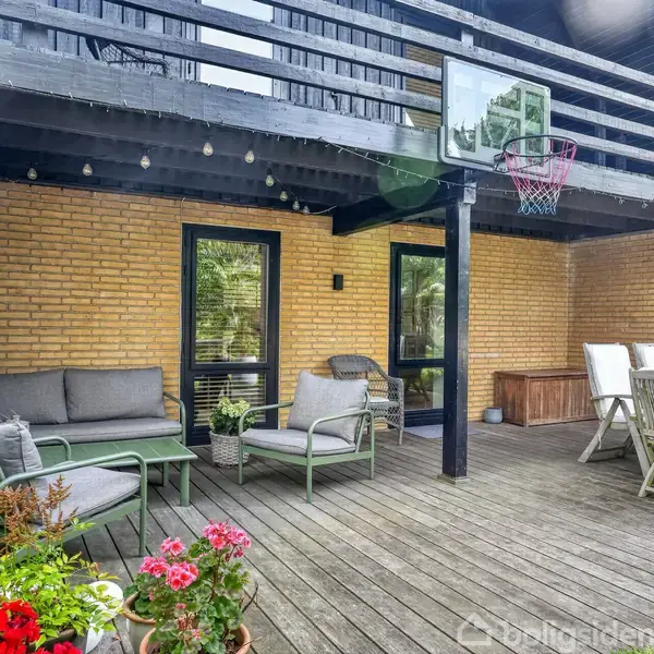 Terrasse med træmøbler; sofa og stole til venstre, spisebord til højre. Omgivelserne inkluderer murstensvægge, planter og en basketballkurv på en træoverdækning.