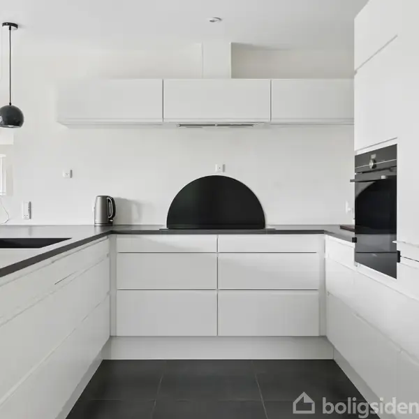 Et moderne, hvidt køkken med sorte bordplader. En indbygget ovn er til højre, og en vask med armatur er til venstre. Rummet er lyst og minimalistisk indrettet.